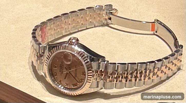 A close-up shot of a two-tone Rolex watch with a rose gold dial and Roman numerals. The watch has a Jubilee bracelet, which is a combination of stainless steel and rose gold. The watch is lying on a textured surface, possibly a display cushion.