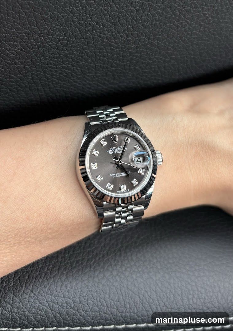 A close-up shot of a Rolex watch on a person's wrist. The watch has a dark grey dial with diamond hour markers and a date window at the 3 o'clock position. The watch is made of stainless steel and features a fluted bezel and a Jubilee bracelet. The background is a dark, textured surface, possibly leather.