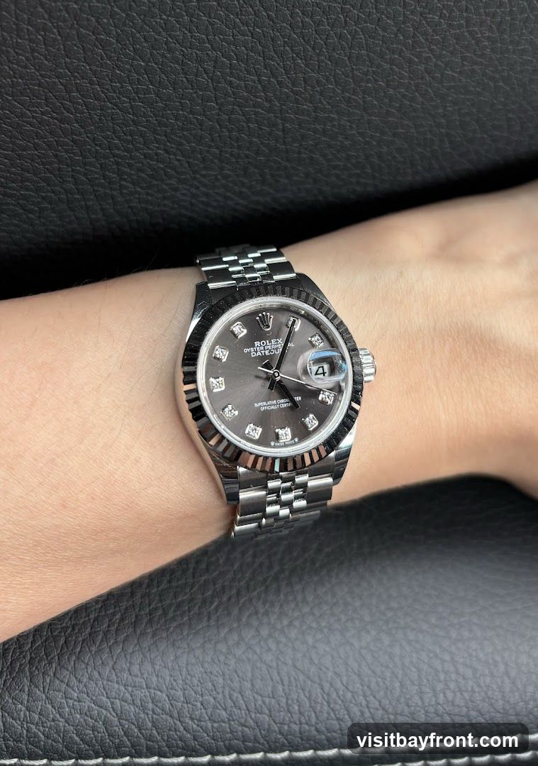 A close-up shot of a Rolex watch on a person's wrist. The watch has a dark grey dial with diamond hour markers and a date window at the 3 o'clock position. The watch is made of stainless steel and features a fluted bezel and a Jubilee bracelet. The background is a dark, textured surface, possibly leather.