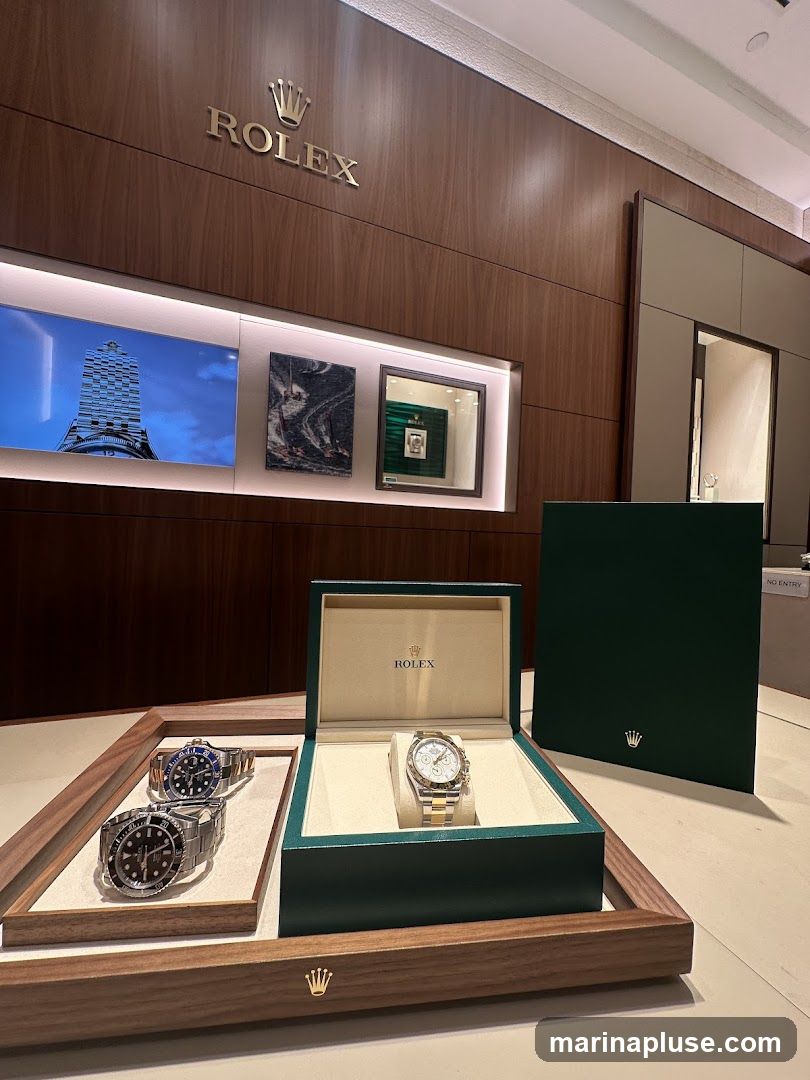 A display of Rolex watches inside a store. The Rolex logo and name are prominently displayed on the wall. Several watches are showcased in open boxes and on trays. One watch is in an open green Rolex box, and two other watches are on a wooden tray. A green Rolex box is also standing upright to the right. Digital screens on the wall show images related to Rolex.