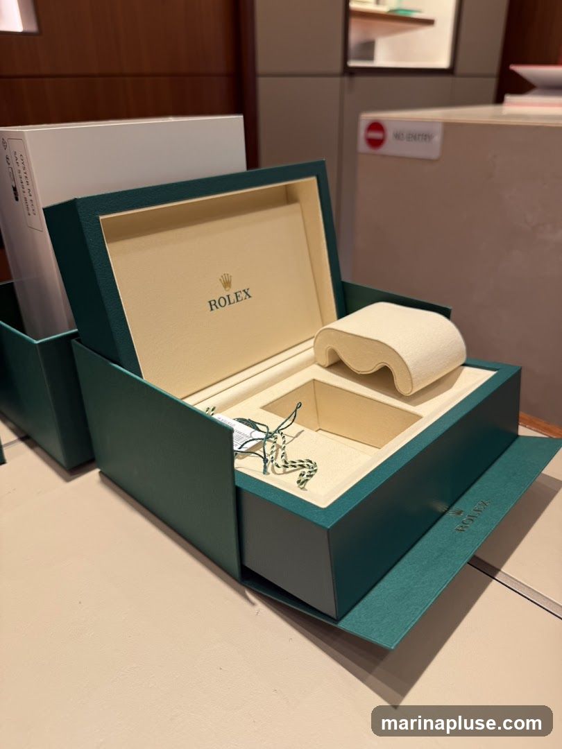 An open Rolex watch box with the brand's logo visible inside. The box is dark green with a cream-colored interior. There is a white pillow for a watch and compartments for accessories. A tag is attached to the box with green and white string. In the background, a sign reads "NO ENTRY".