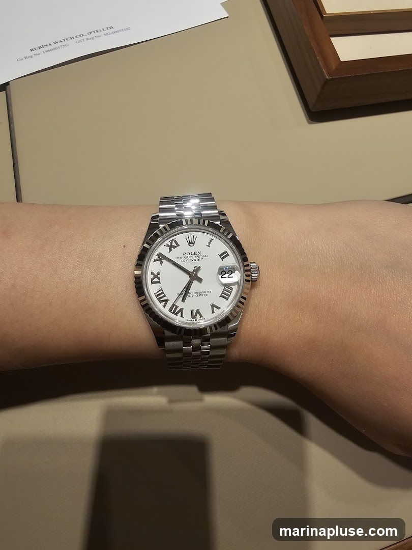 A close-up shot of a person's wrist wearing a Rolex Oyster Perpetual Datejust watch. The watch has a white dial with Roman numerals and a date window at the 3 o'clock position. The watch band is a Jubilee bracelet. In the background, a document with the text "RUBINA WATCH CO. (PTE) LTD." is visible.
