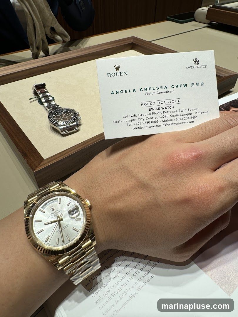 A close-up shot of two Rolex watches, one on a person's wrist and another displayed in a wooden tray. A business card for Rolex Boutique, featuring the name Angela Chelsea Chew, Watch Consultant, and contact details for the Kuala Lumpur store, is also visible. The background shows a retail environment with a person wearing gloves, likely a sales associate.