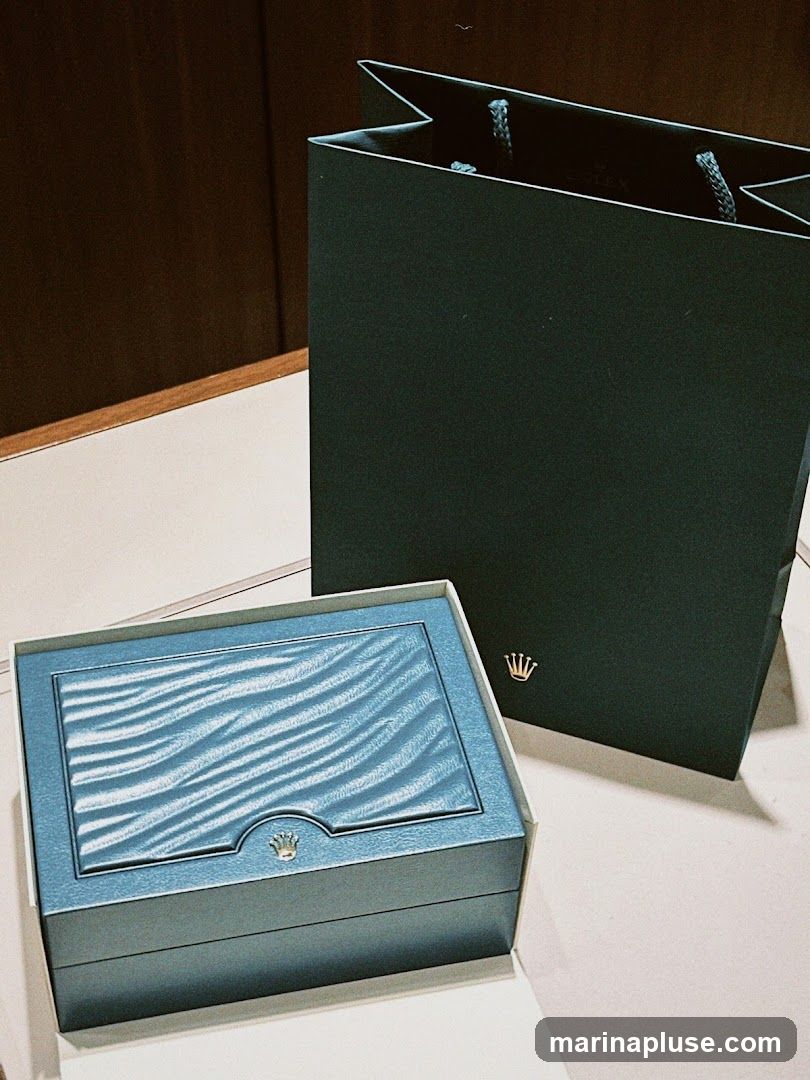 A teal blue Rolex watch box with a textured lid and a small crown logo is placed on a white surface. Behind it is a dark green Rolex shopping bag with rope handles and a gold crown logo. The background is a dark wood panel.