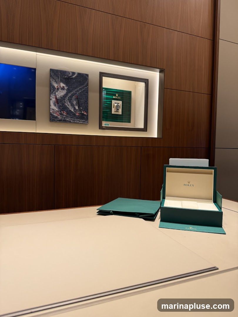A display case with a Rolex watch inside, next to artwork of sailboats. On the counter, there is a green Rolex shopping bag and an open Rolex watch box.