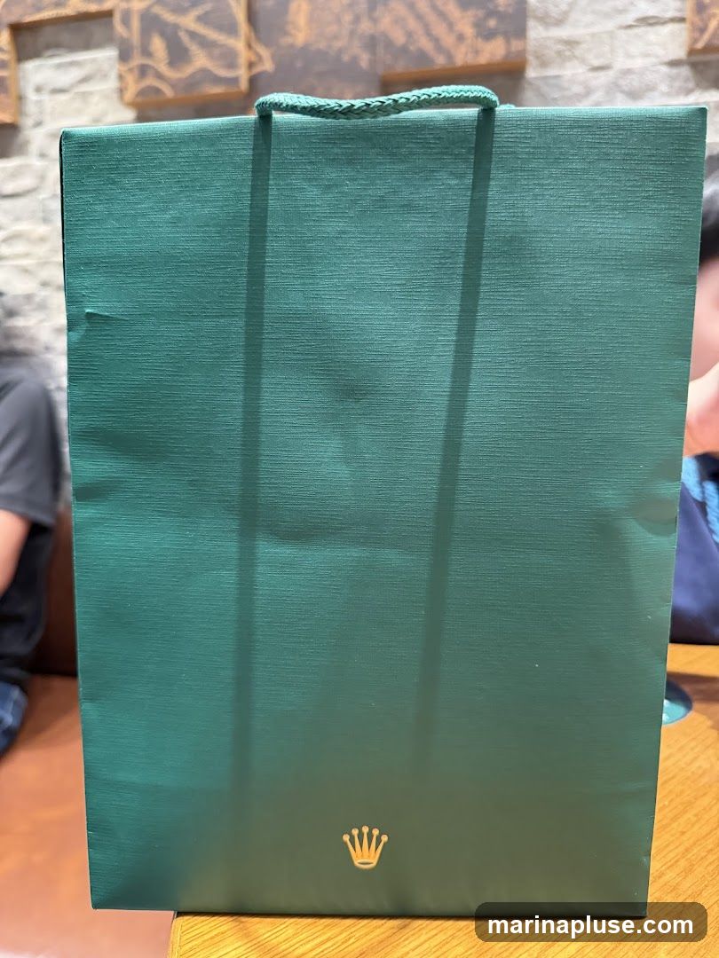 A green paper gift bag with braided handles is shown. The Rolex crown logo is printed in gold at the bottom center of the bag. The bag is placed on a wooden surface, and parts of people are visible in the background.