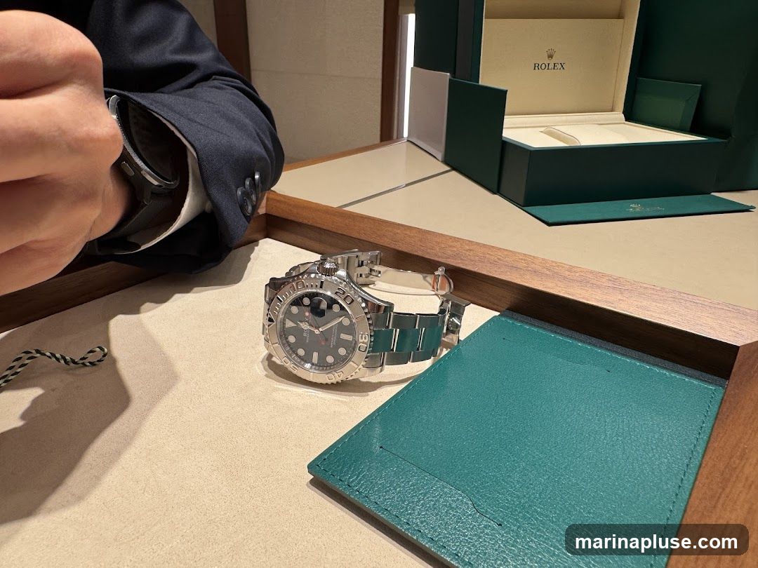 A person in a dark suit is trying on a silver Rolex watch. The watch is displayed on a light-colored surface with a green leather insert. In the background, an open Rolex watch box is visible.
