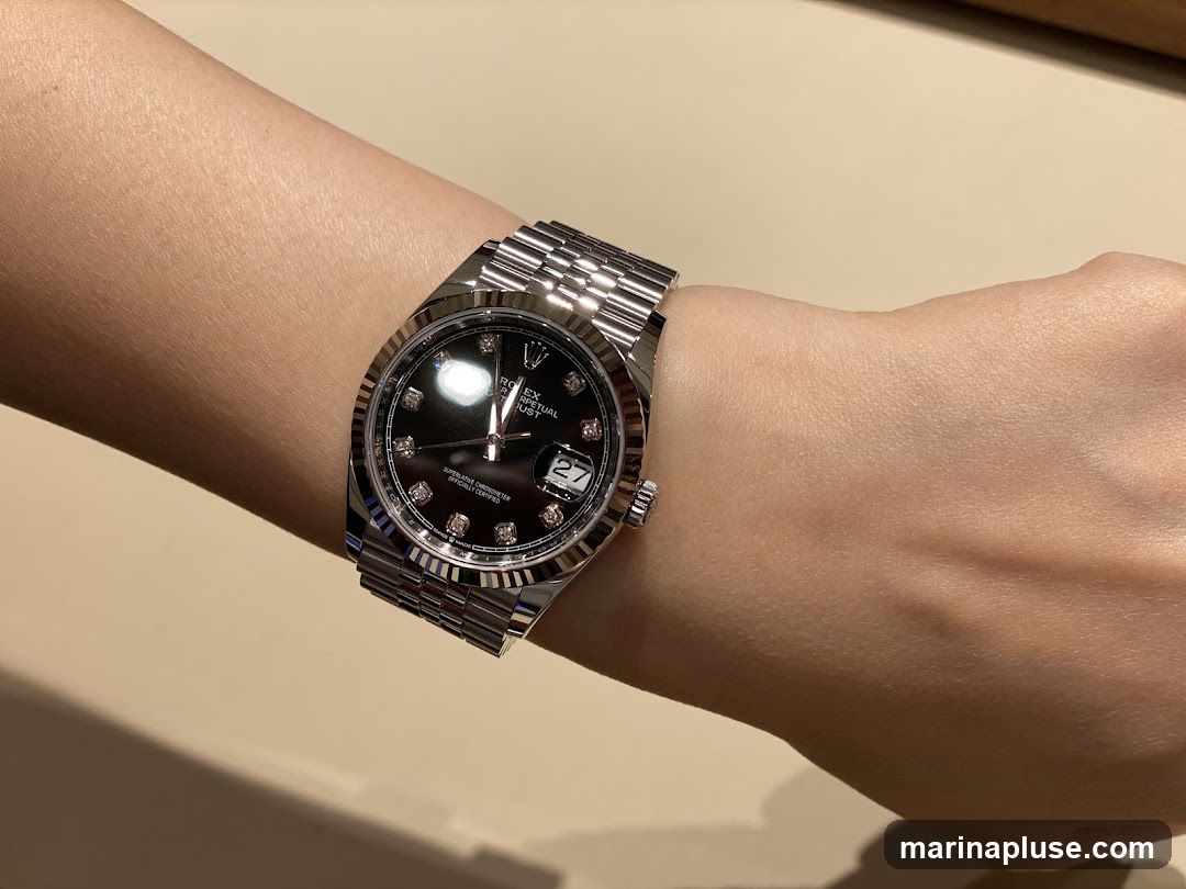 A close-up shot of a person's wrist wearing a silver Rolex watch with a black dial and diamond hour markers. The watch has a fluted bezel and a Jubilee bracelet. The date window shows '27'.