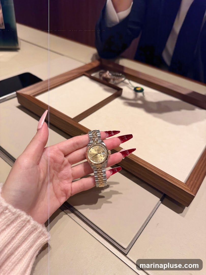 A woman's hand with long, red-tipped nails holds a two-tone Rolex watch. The watch has a gold dial with diamond markers and a two-tone oyster bracelet. The background shows a display case with other jewelry and a person in a suit.