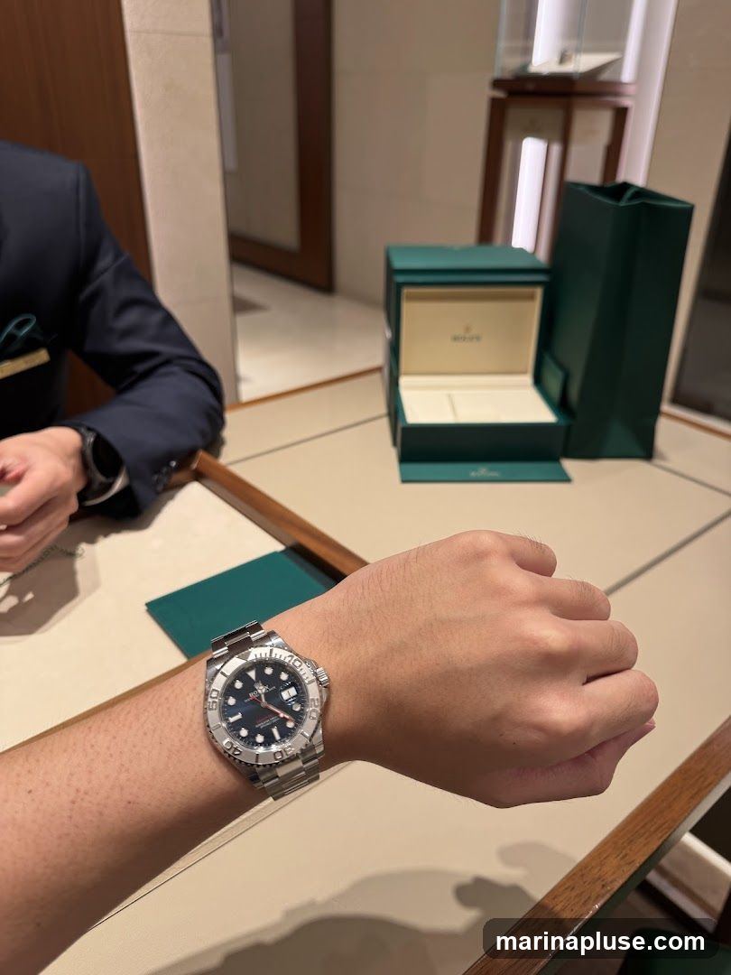 A person is wearing a Rolex watch on their wrist. In the background, a Rolex watch box and shopping bag are displayed on a table. The scene appears to be in a luxury watch store.