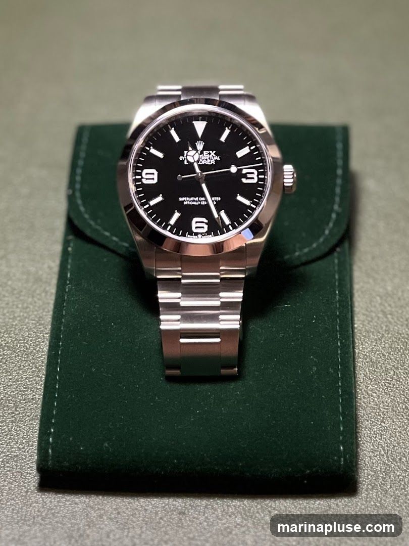 A close-up shot of a Rolex Explorer watch with a black dial and stainless steel bracelet, resting on a dark green velvet pouch. The watch face displays the Rolex logo, "Oyster Perpetual Explorer," and "Superlative Chronometer Officially Certified." The numbers 3, 6, and 9 are visible on the dial.