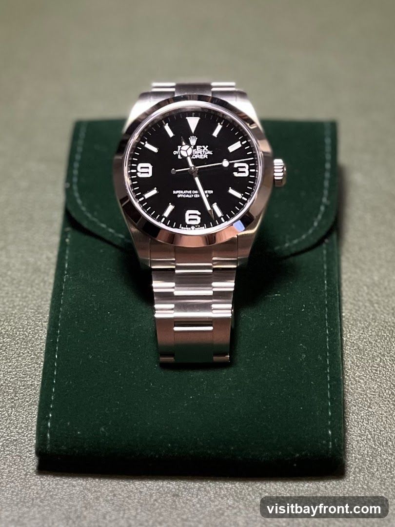 A close-up shot of a Rolex Explorer watch with a black dial and stainless steel bracelet, resting on a dark green velvet pouch. The watch face displays the Rolex logo, "Oyster Perpetual Explorer," and "Superlative Chronometer Officially Certified." The numbers 3, 6, and 9 are visible on the dial.