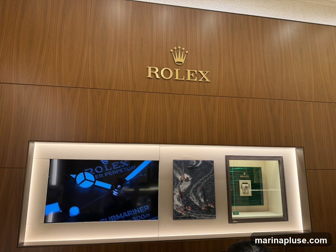 A Rolex logo and crown are displayed prominently on a wood-paneled wall. Below the logo, a digital display shows a close-up of a Rolex watch face with blue luminous hands and markers, and text indicating "ROLEX OYSTER PERPETUAL SUBMARINER 300m". To the right of the screen, a framed artwork depicts sailboats on a choppy sea. Further to the right, a Rolex watch is showcased in a glass display case with a green background.