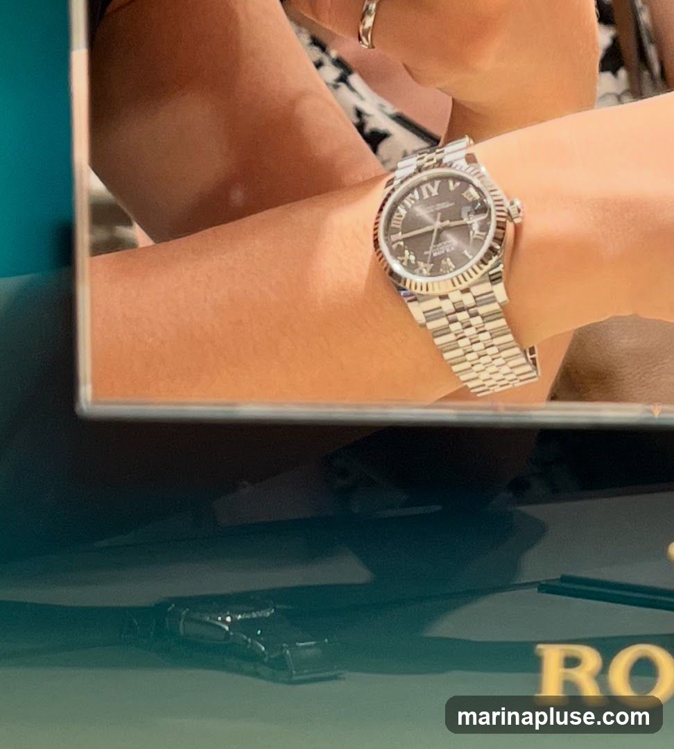 A close-up shot shows a person's wrist wearing a silver watch with a dark face and Roman numerals. The watch band is made of interlocking metal links. In the reflection below, the word "ROLEX" is visible in gold lettering, along with a small black car model.
