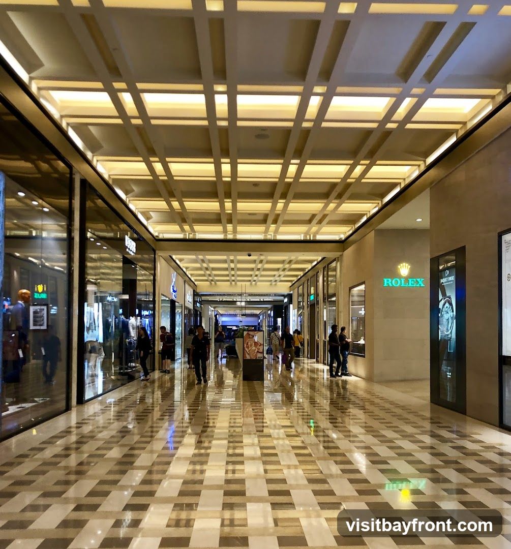 A view down a well-lit, modern shopping mall corridor with a high, coffered ceiling. Shops line both sides of the walkway, with large glass windows displaying merchandise. The floor is polished marble with a geometric pattern, reflecting the lights and the ceiling structure. Several people are walking through the mall. A prominent ROLEX store is visible on the right side.