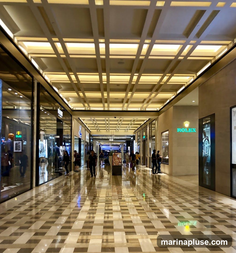 A view down a well-lit, modern shopping mall corridor with a high, coffered ceiling. Shops line both sides of the walkway, with large glass windows displaying merchandise. The floor is polished marble with a geometric pattern, reflecting the lights and the ceiling structure. Several people are walking through the mall. A prominent ROLEX store is visible on the right side.