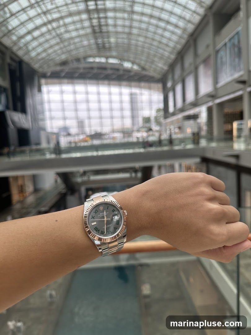 A person's wrist wearing a two-tone Rolex watch is shown in the foreground. The background is a blurred view of a large, modern indoor shopping mall with a glass ceiling and multiple levels.