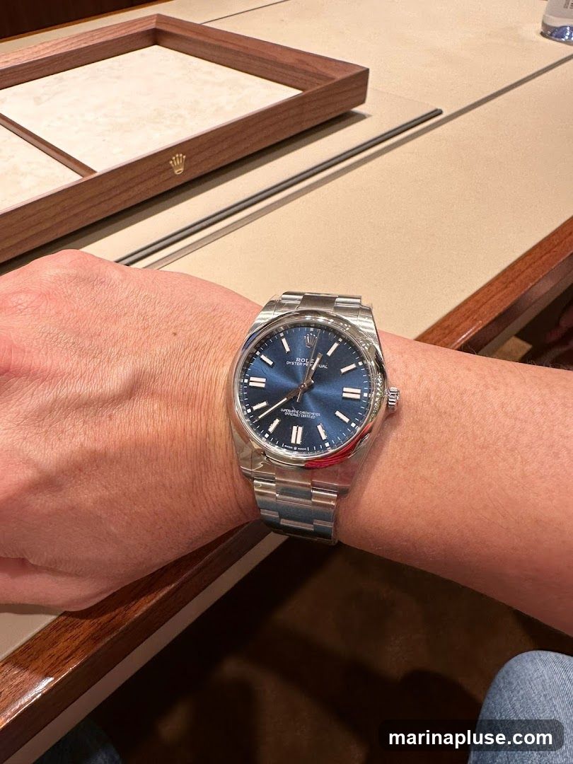 A person's wrist wearing a silver Rolex watch with a blue dial. The watch is on a metal band. In the background, a wooden display tray with a gold Rolex crown logo is visible. The setting appears to be a jewelry store or watch boutique.