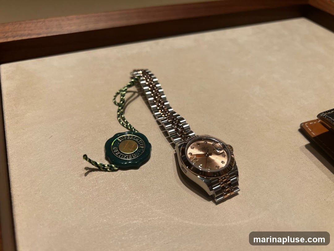 A close-up shot of a rose gold and stainless steel Rolex watch with a champagne dial, displayed on a beige surface. A green circular certification tag with gold lettering hangs from the watch by a green string. The tag reads "SUPERLATIVE CHRONOMETER OFFICIALLY CERTIFIED". Part of another watch strap or accessory is visible on the right side of the image.