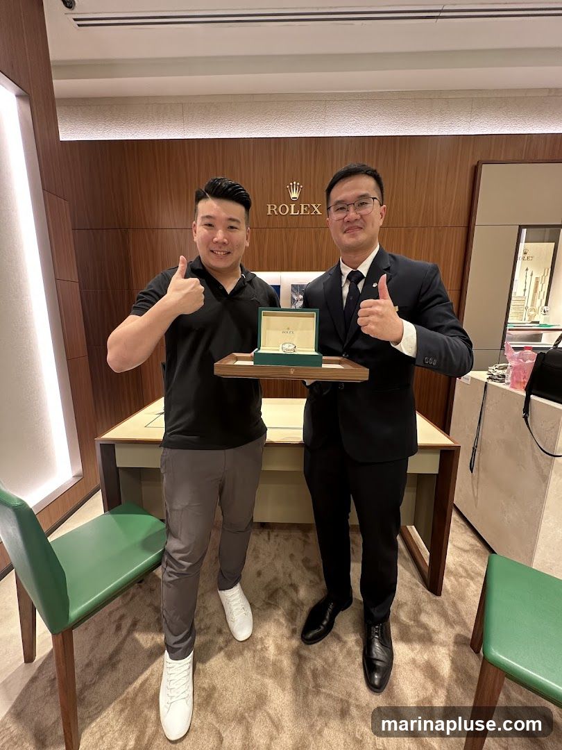Two men are standing in a store, holding a Rolex watch box. The man on the left is wearing a black polo shirt and grey pants, and is giving a thumbs up. The man on the right is wearing a suit and tie, and is also giving a thumbs up. The Rolex logo is visible on the wall behind them. There are green chairs in the foreground and a display case in the background.