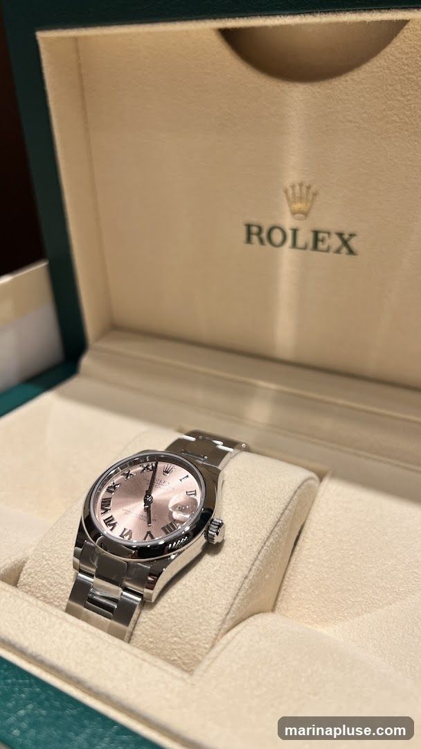 A close-up shot of a Rolex watch with a pink dial and Roman numerals, resting in its presentation box. The Rolex logo and name are visible on the inside lid of the box.