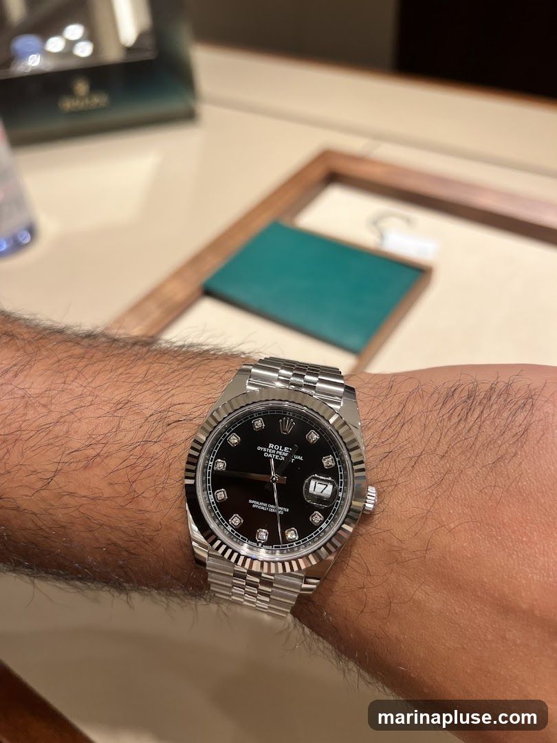 A close-up shot of a person's wrist wearing a silver Rolex watch with a black dial and diamond hour markers. The watch has a fluted bezel and a Jubilee bracelet. In the background, a blurred green box with the Rolex logo is visible, along with a display case.