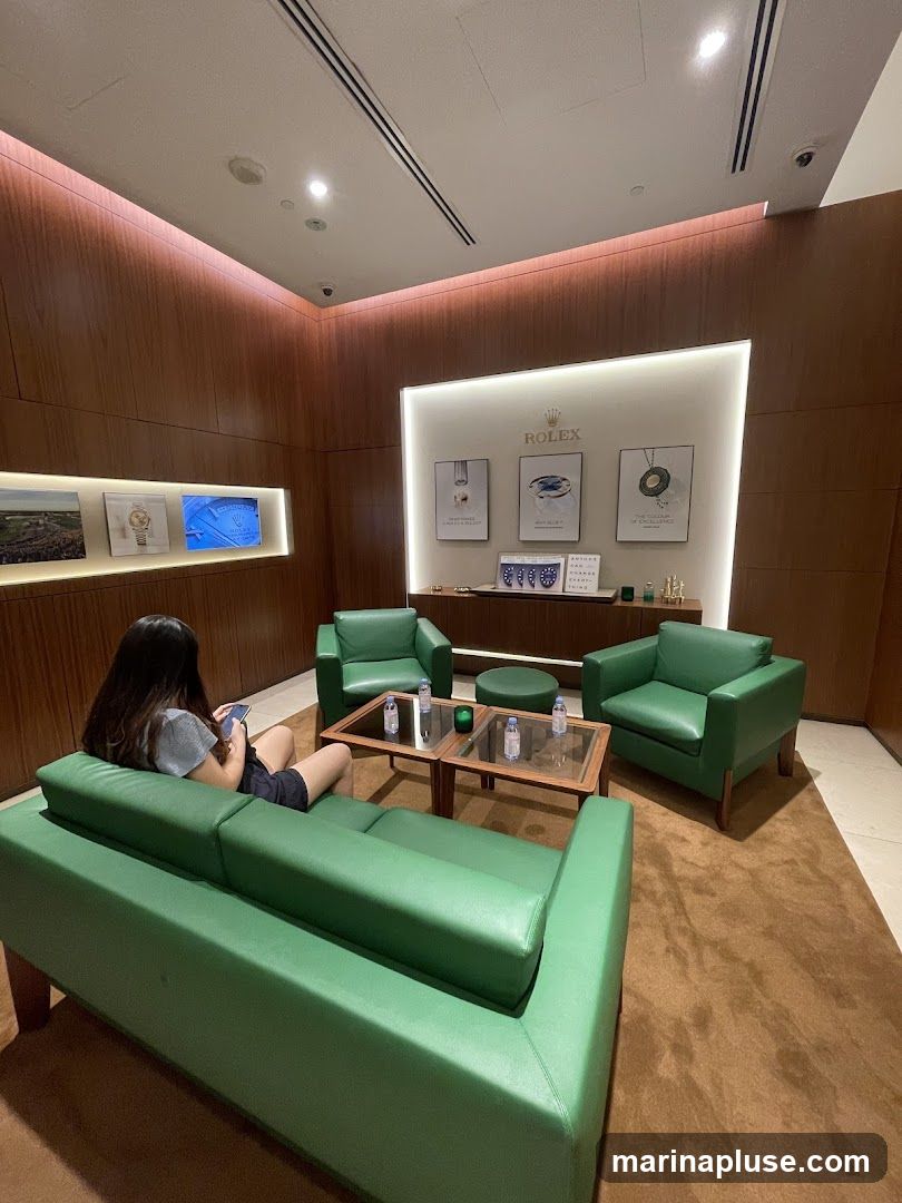 A woman sits on a green sofa in a Rolex showroom, looking at her phone. The room features wood-paneled walls, a display of Rolex watches and artwork, and green armchairs around coffee tables with bottled water.