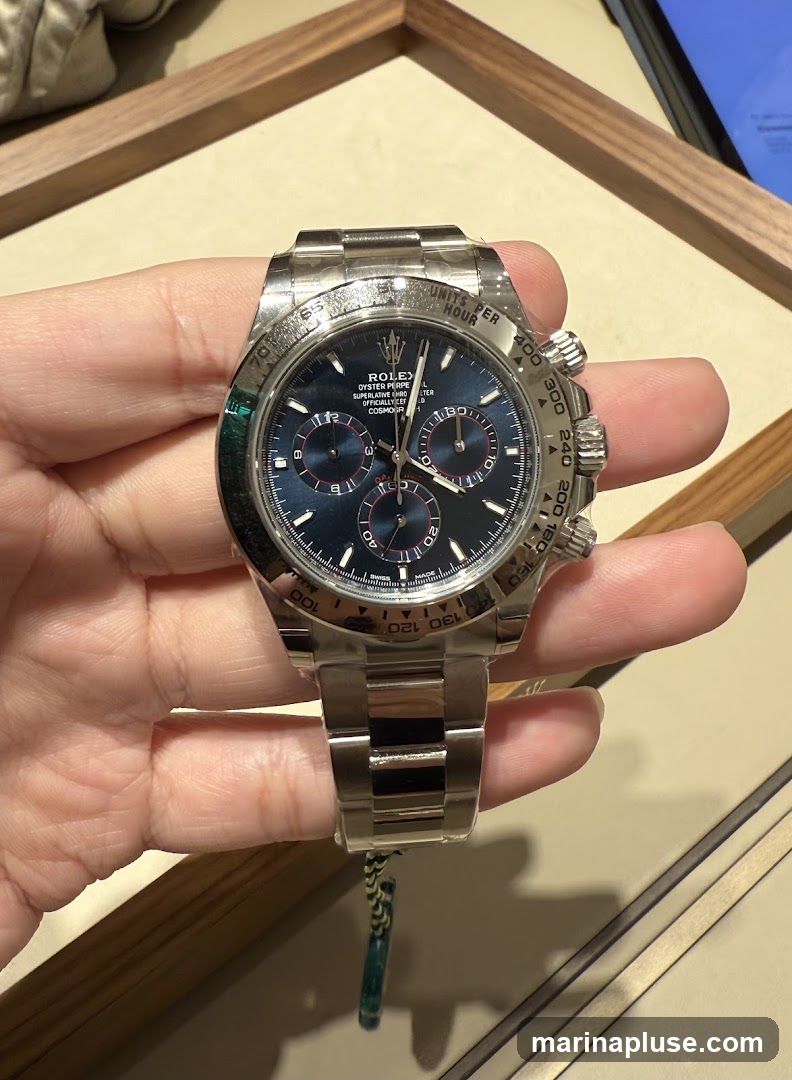 A person is holding a Rolex Cosmograph Daytona watch with a blue dial. The watch has a stainless steel band and a tachymeter bezel. The dial features three sub-dials and luminous hour markers. The watch is displayed against a blurred background with wooden accents.