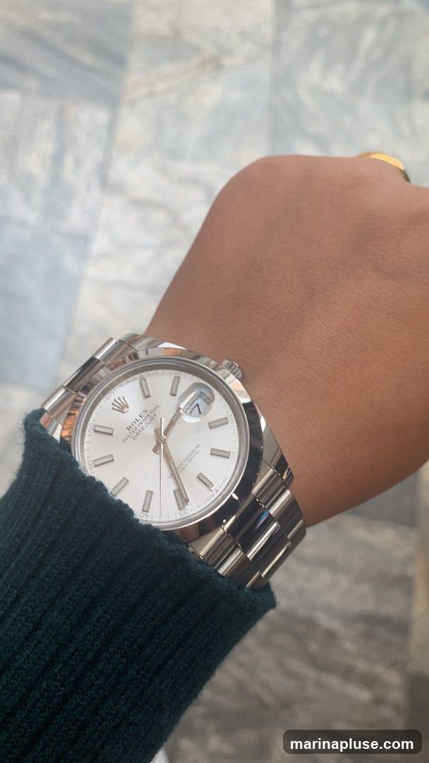 A close-up shot of a person's wrist wearing a silver Rolex Oyster Perpetual Datejust watch. The watch has a white dial with silver hour markers and hands, and a date window at the 3 o'clock position. The watch is partially covered by a dark green sweater sleeve. A gold ring is visible on the person's ring finger. The background appears to be a marble surface.