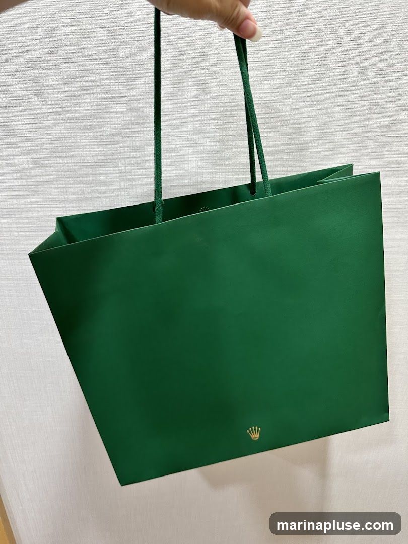 A hand holds a green paper shopping bag with green rope handles. A small gold Rolex crown logo is visible on the lower right side of the bag. The background is a textured white wall.