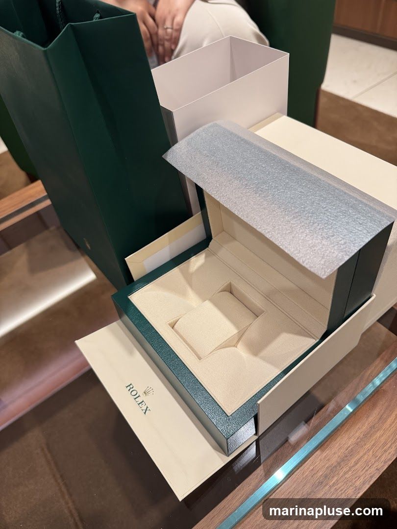 Several Rolex watch boxes and a shopping bag are arranged on a table. The boxes are open, revealing the interior compartments designed to hold watches. A person's hands are visible in the background, wearing a ring. The Rolex logo is visible on a piece of paper and on the shopping bag.