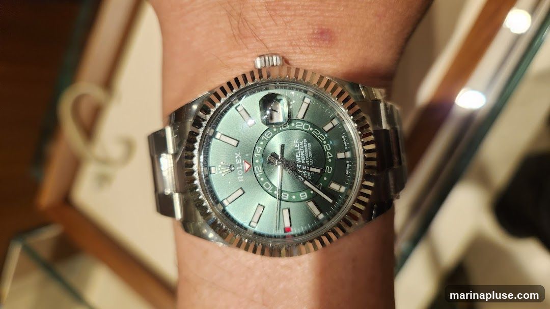 A close-up shot of a Rolex Sky-Dweller watch on a person's wrist. The watch has a green dial with white hour markers and a fluted bezel. The dial displays the time, date, and a second time zone. The background is blurred, showing what appears to be a display case or box.