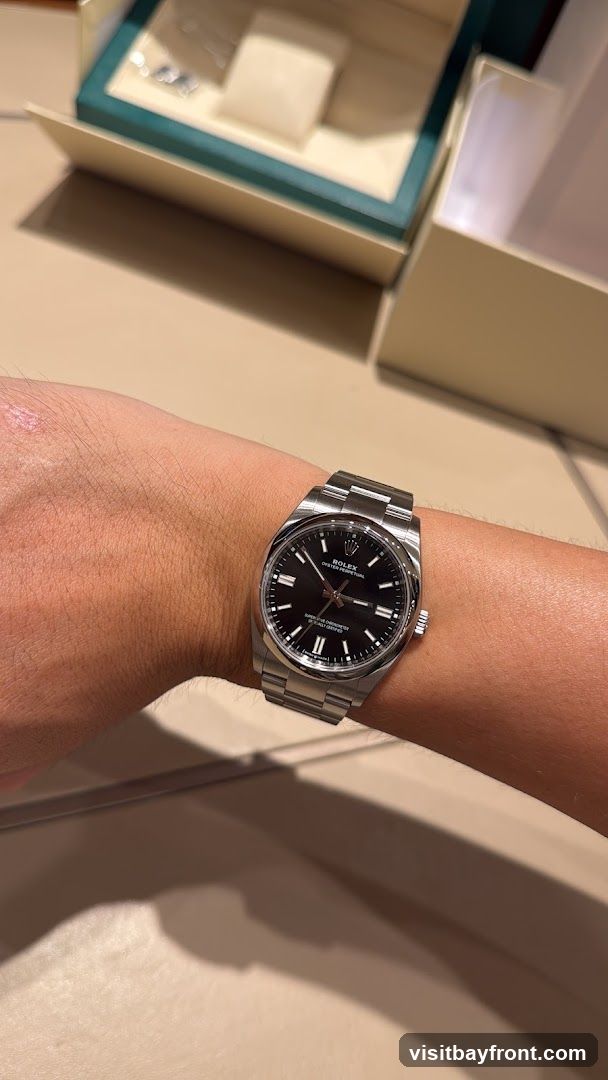 A close-up shot of a person's wrist wearing a Rolex Oyster Perpetual watch with a black dial. The watch has a stainless steel band and case. In the background, a Rolex watch box is partially visible.