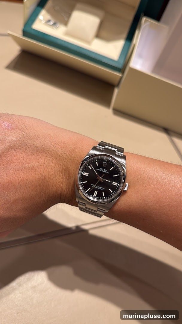 A close-up shot of a person's wrist wearing a Rolex Oyster Perpetual watch with a black dial. The watch has a stainless steel band and case. In the background, a Rolex watch box is partially visible.