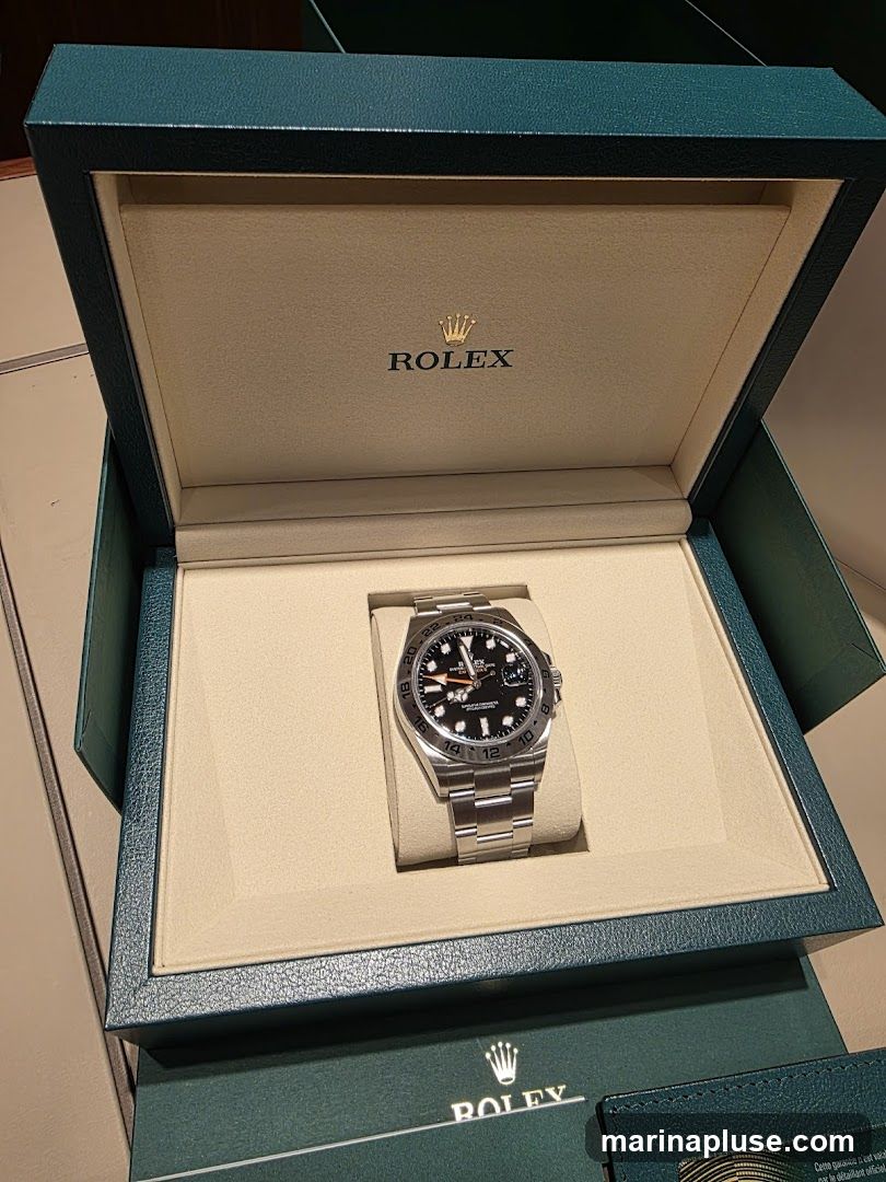 A silver Rolex watch with a black dial is displayed inside an open, teal-colored presentation box. The inside of the box is lined with cream-colored material and features the Rolex logo and crown in gold. Another Rolex branded box or sleeve is visible at the bottom of the image.