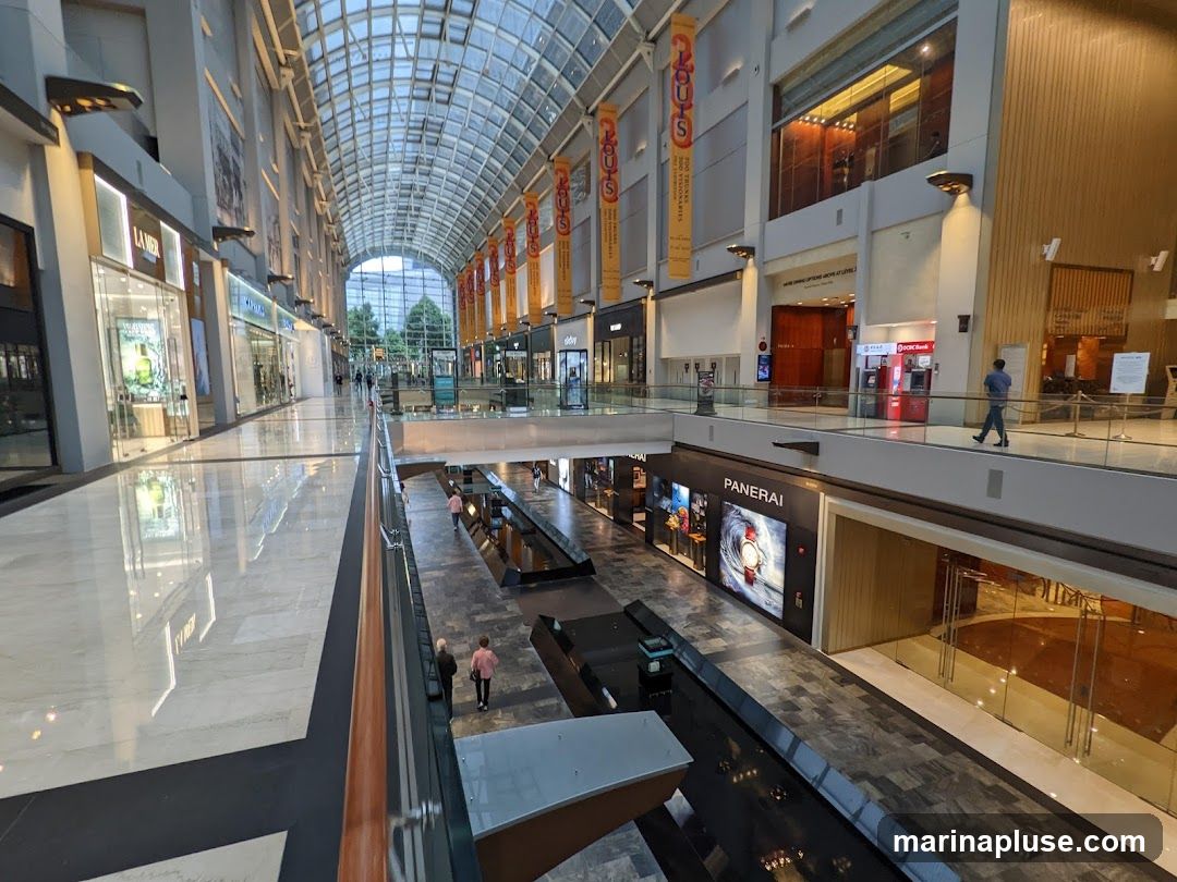 A wide-angle view of the interior of a large, modern shopping mall. The mall features a high, arched glass ceiling that allows natural light to filter in. On the left, several storefronts are visible, including one with a sign for 'LA MER'. On the right, a walkway overlooks a lower level where more shops are located, including a prominent display for 'PANERAI' watches. People are seen walking on both levels. The architecture is sleek and contemporary, with polished floors and clean lines. Banners hang from the upper level, displaying text that is mostly illegible.