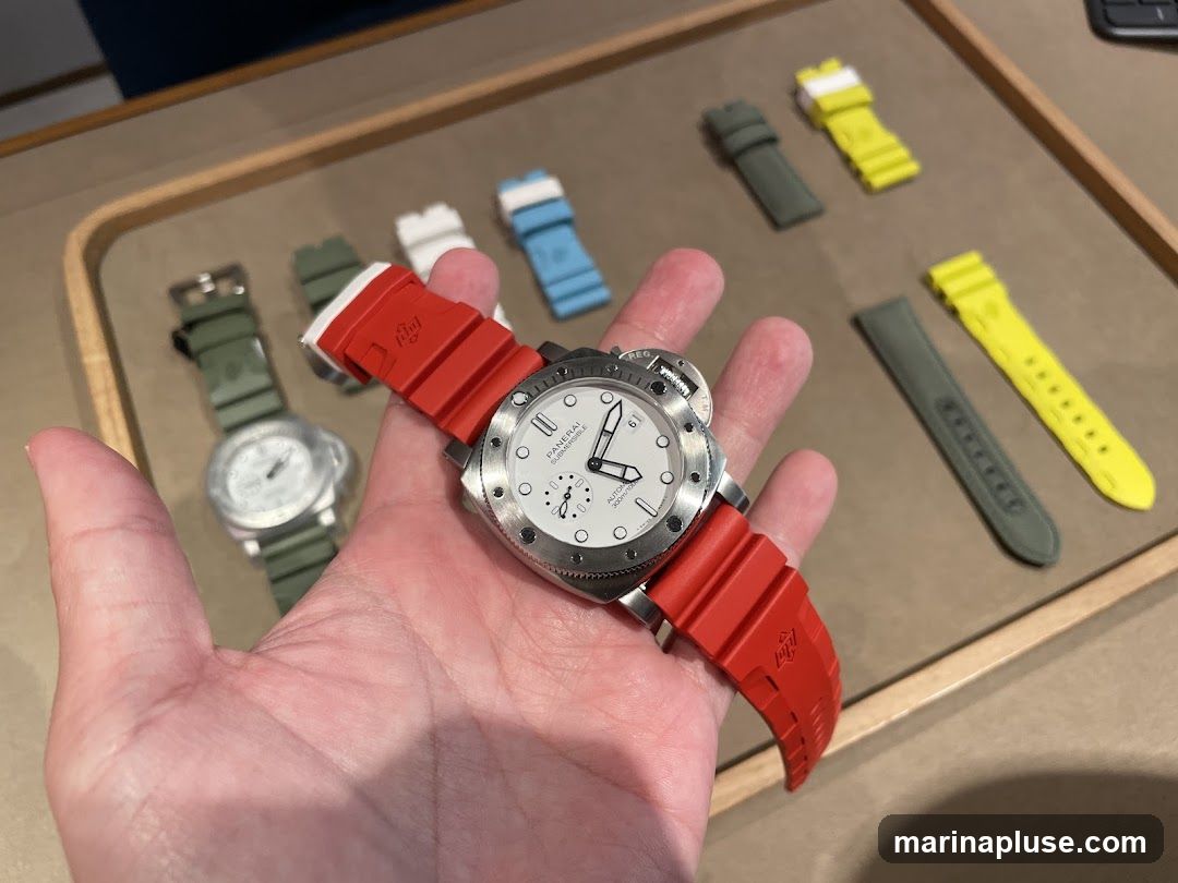 A hand holds a silver watch with a red rubber strap. In the background, on a wooden tray, are other watches and various colored watch straps, including green, white, blue, and yellow.