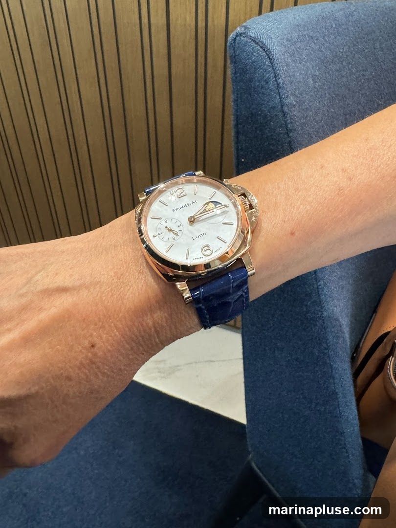 A close-up shot of a person's wrist wearing a rose gold Panerai watch with a white dial and a blue alligator leather strap. The watch face shows the brand name "PANERAI" and "Luna" along with a moon phase complication. The background features a textured wooden wall and a blue upholstered chair.