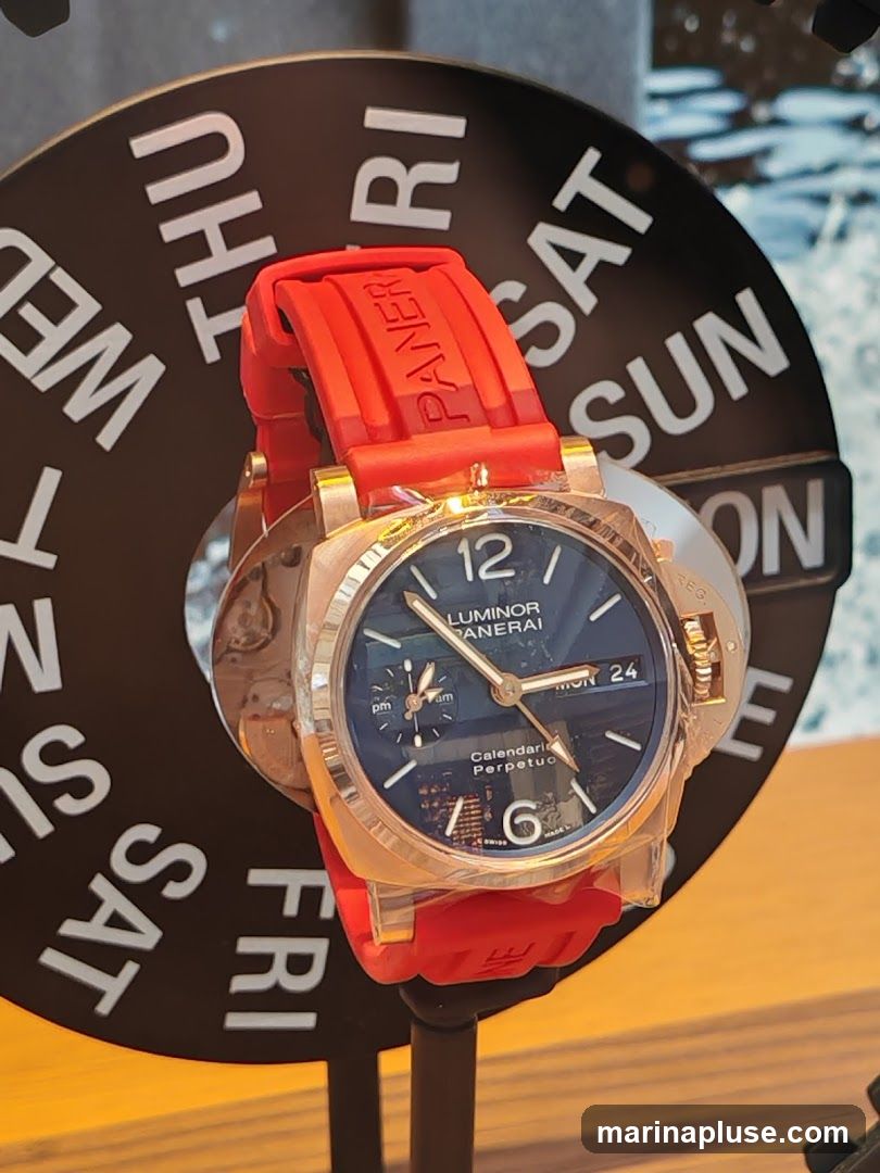 A close-up of a Panerai Luminor Perpetual Calendar watch with a blue dial and a red rubber strap. The watch is displayed on a stand in front of a black circular display with days of the week printed in white. The days visible are Wednesday, Thursday, Friday, Saturday, and Sunday. The watch face shows the time, date (Monday 24), and sub-dials for AM/PM and perpetual calendar functions.