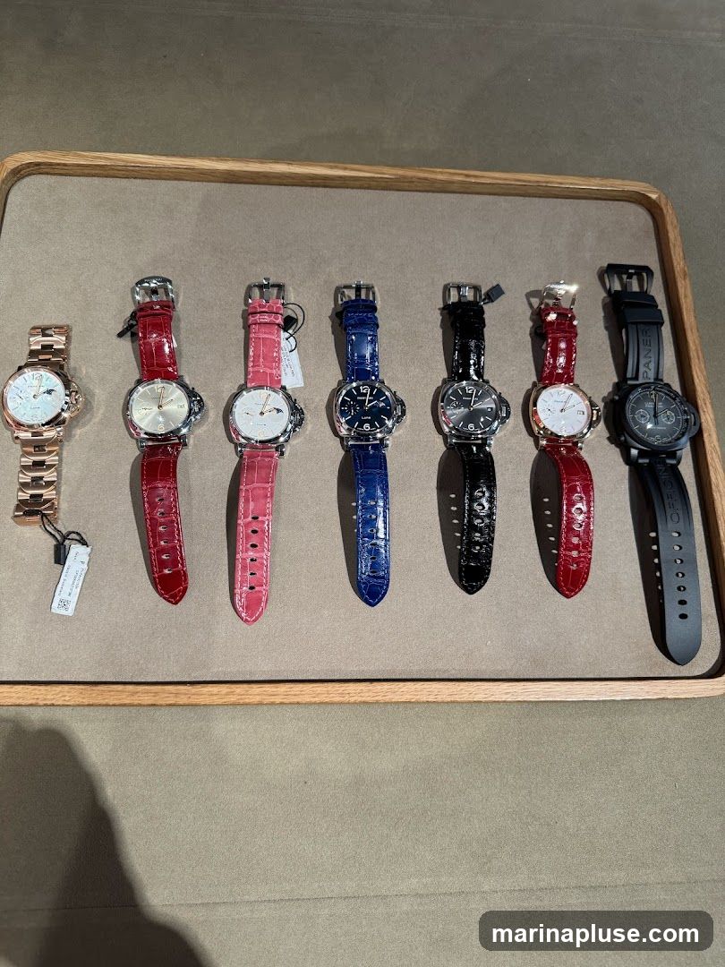 A display of seven luxury watches with various colored leather straps and metal bands, laid out on a neutral-colored surface within a wooden frame. The watches feature different dial designs and case materials.