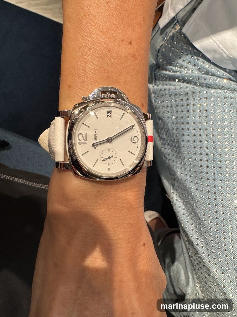 A close-up shot of a person's wrist wearing a Panerai watch. The watch has a white dial with black Arabic numerals and a date window at the 3 o'clock position. The watch band is white with red and blue accents. The person is wearing a white shirt and denim jeans adorned with rhinestones.