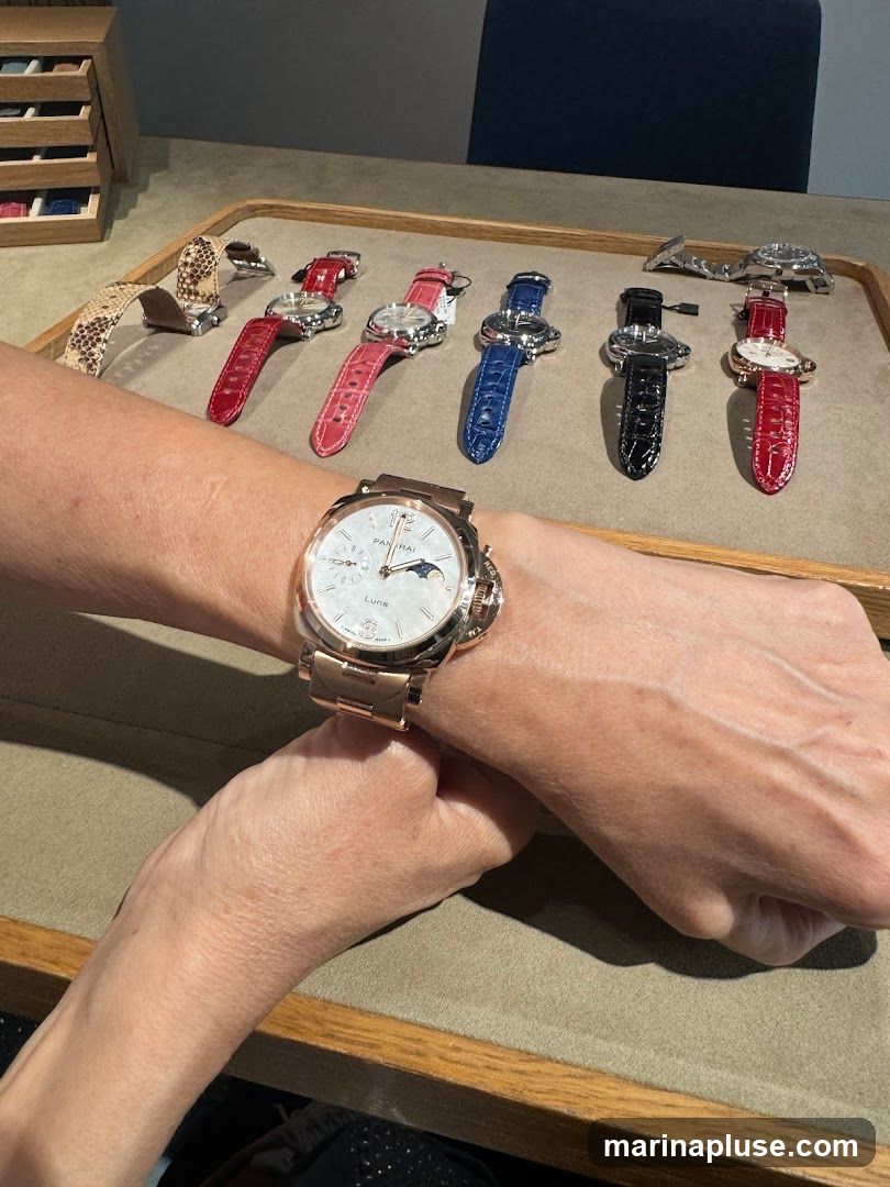 A person is wearing a rose gold wristwatch with a white dial. In the background, several other wristwatches with colorful straps (red, pink, blue, black, and snakeskin) are displayed on a tray. A wooden organizer with watch straps is visible in the upper left corner.