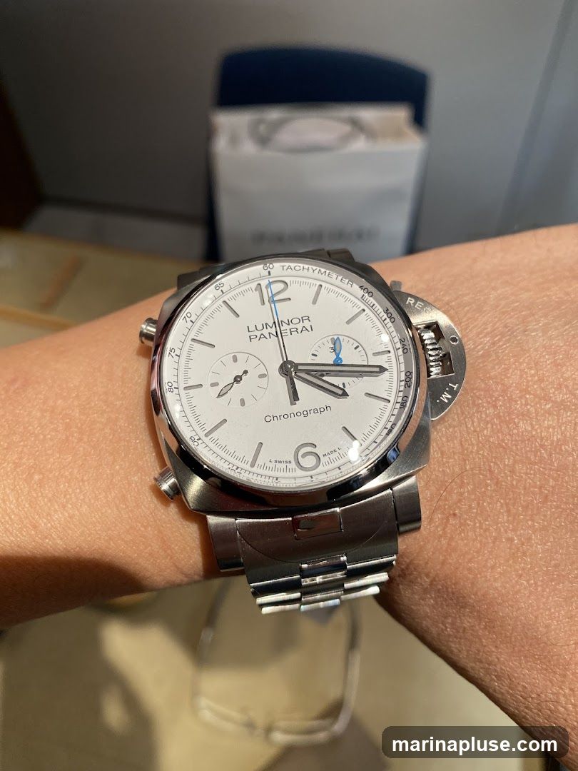A close-up shot of a Panerai Luminor Chronograph watch on a person's wrist. The watch has a white dial with silver hands and markers. The dial features a tachymeter scale around the edge and two subdials. The watch is made of stainless steel and has a metal bracelet. In the background, a blurred white box with text is visible.