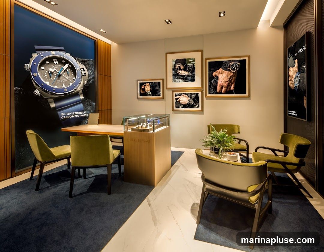A well-lit and modern watch store interior featuring a large wall graphic of a Panerai Submersible Chronograph watch. Several framed photographs of divers and watches are displayed on the wall. A wooden counter with a glass display case showcasing watches is centrally located. Seating areas with plush green chairs are arranged around the space. A vertical display panel with Panerai branding is visible on the right wall.