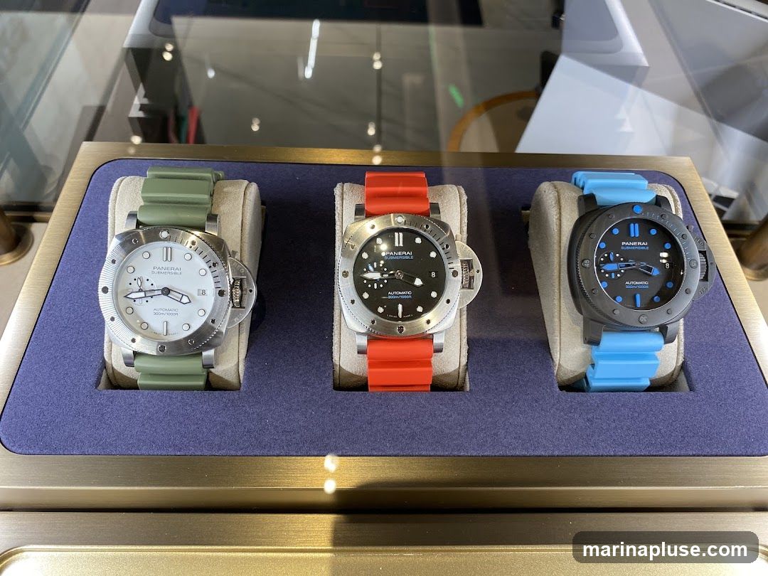 Three Panerai Submersible watches are displayed in a glass case. The watches have white, black, and dark grey faces, respectively, and are fitted with green, orange, and blue rubber straps. The watch on the left has a white face with black markers and text. The middle watch has a black face with white markers and text. The watch on the right has a dark grey face with blue markers and text. All watches are automatic and water-resistant to 300m/1000ft.