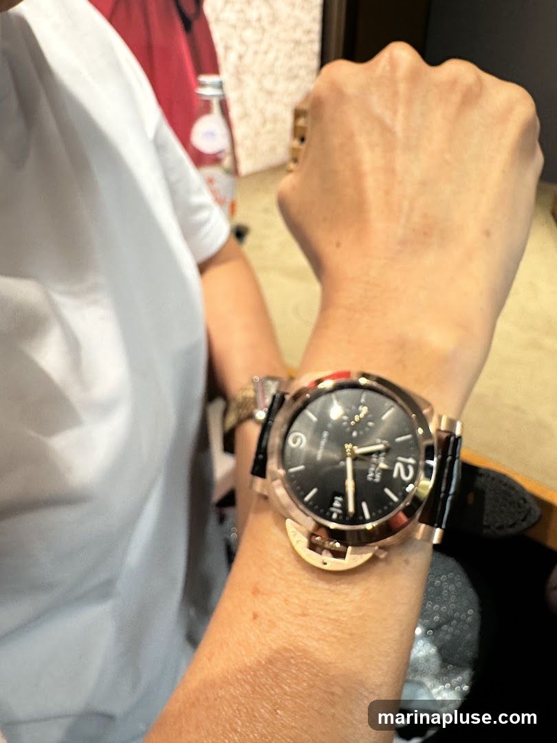 A close-up shot of a person's arm wearing a rose gold watch with a black dial and strap. The watch has large Arabic numerals for the hour markers, with the number 12 prominently displayed. The background is blurred, showing hints of a retail environment with a bottle and some display items.