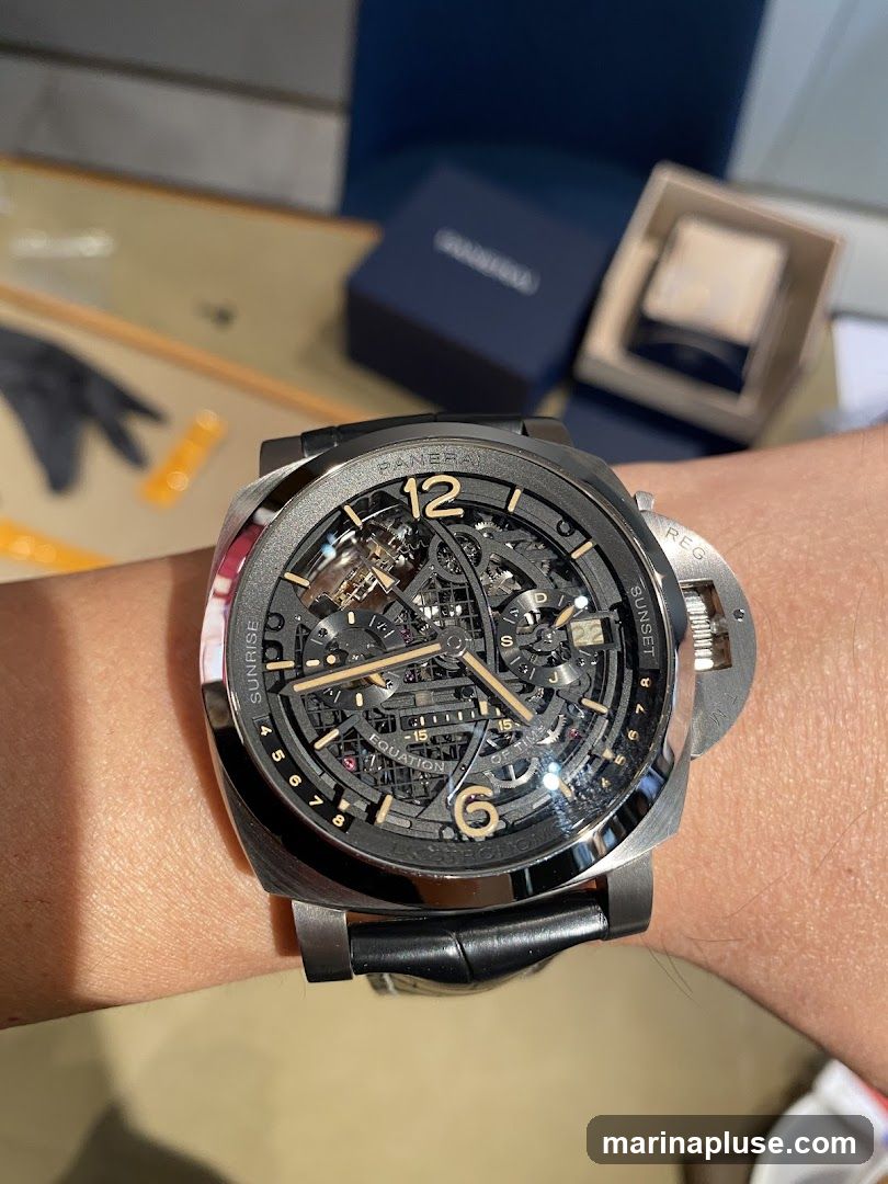 A close-up shot of a Panerai skeleton watch on a person's wrist. The watch features a complex mechanical movement visible through the dial, with gold-toned hands and hour markers. The bezel is dark gray, and the strap is black leather. In the background, a blue Panerai watch box and another watch box are visible.