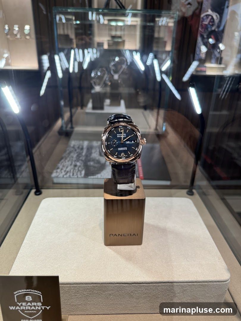 A close-up view of a Panerai watch displayed in a glass case. The watch has a dark blue dial with rose gold accents and a brown leather strap. It is presented on a rose gold stand with the Panerai logo. In the background, other watches and display items are visible within illuminated glass cabinets. A card indicating an '8 Years Warranty' is placed in the foreground.