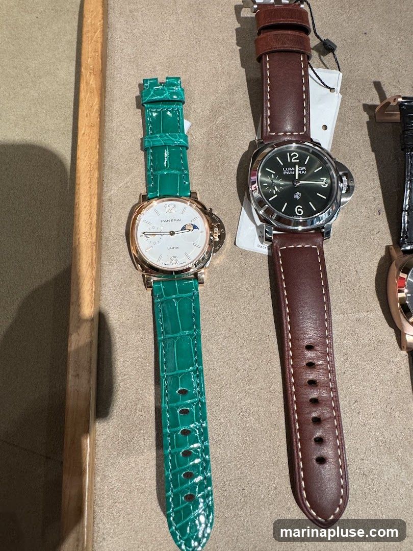 A close-up shot of two luxury watches. The watch on the left has a white face with a moon phase complication and a bright green alligator leather strap. The watch on the right has a dark green face with luminous hour markers and a brown leather strap. Both watches appear to be from the Panerai brand.