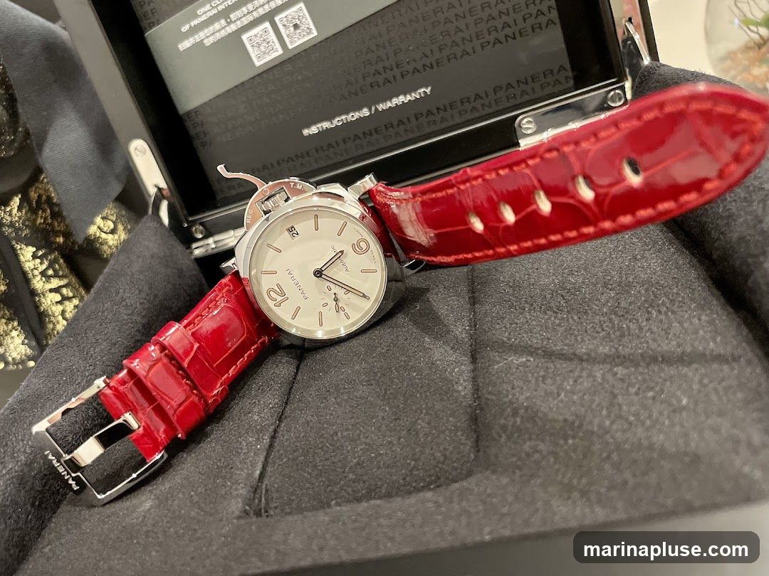 A close-up of a Panerai watch with a white dial and a red alligator leather strap. The watch is in its box, which is open, revealing instructions and warranty information. The watch face shows the time with Arabic numerals for 12 and 6, and a date window at 3 o'clock. The brand name "PANERAI" is visible on the dial and the buckle.